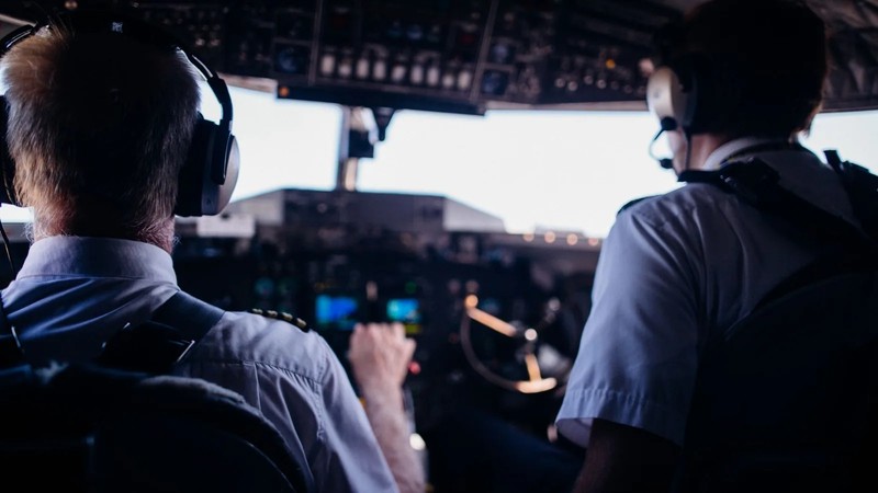 A close-up of a pilot and co-pilot in a cockpit, with an axe visible on the floor.