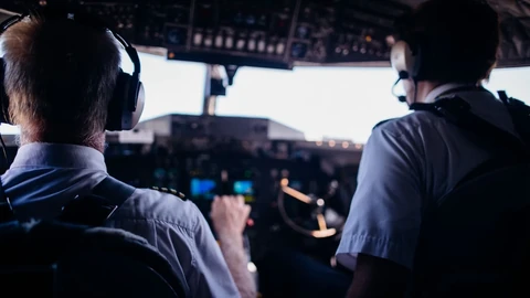 A close-up of a pilot and co-pilot in a cockpit, with an axe visible on the floor.